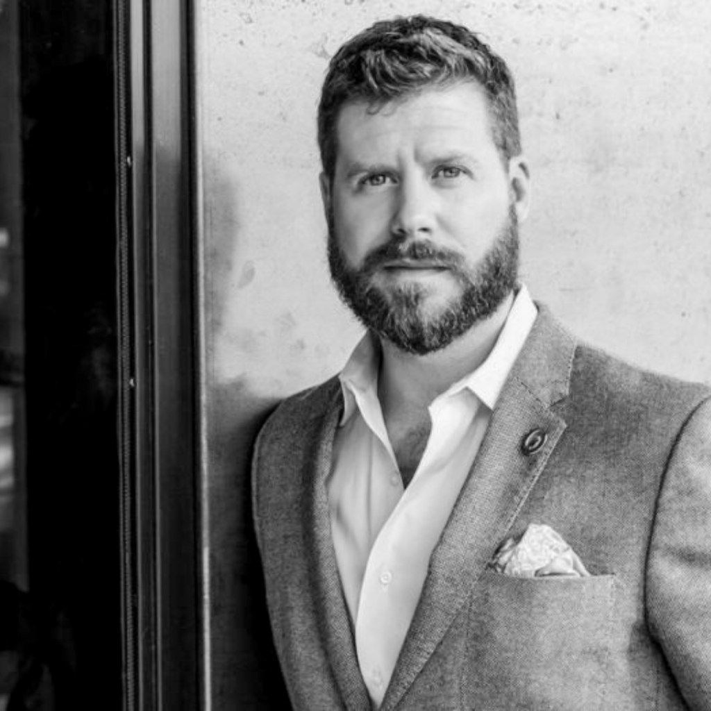 Professional portrait of a man with a beard, wearing a gray blazer and white shirt, standing near a wall.
