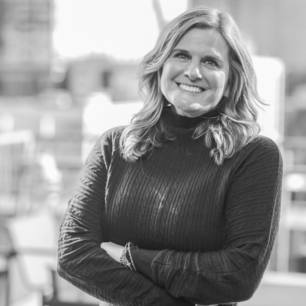 Black and white portrait of a smiling woman with long hair, wearing a black sweater, standing outdoors with arms crossed.