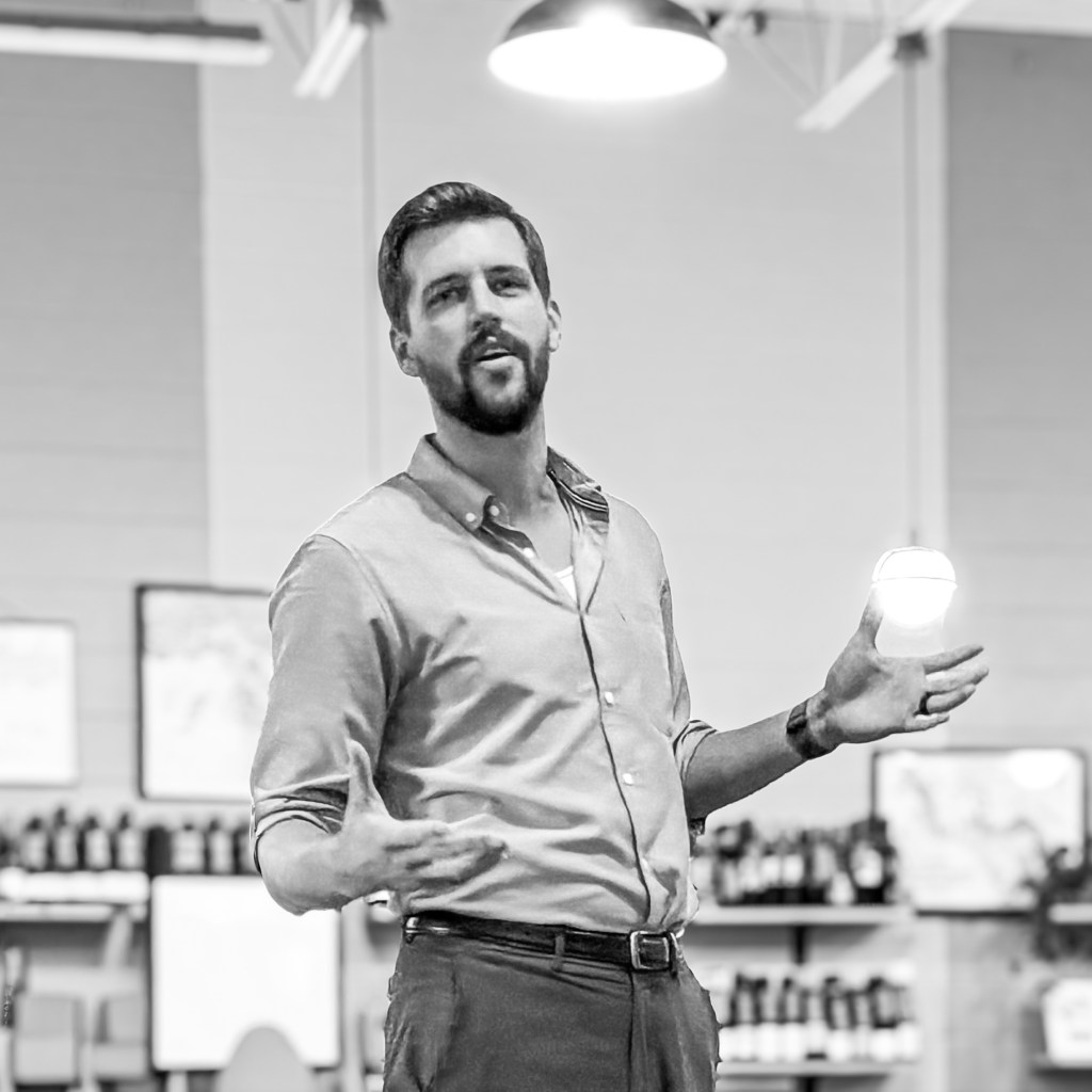 A man in a light shirt gestures while speaking in a professional setting with wine bottles in the background.