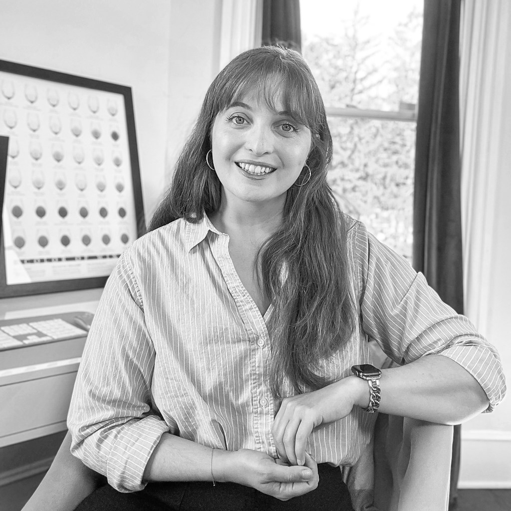 A woman sitting in an office setting, wearing a striped button-up shirt, smiling at the camera, with a framed graphic on the wall behind her.