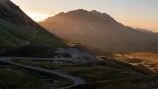 A scenic landscape at sunset featuring rolling hills and a winding road leading towards a mountain range.