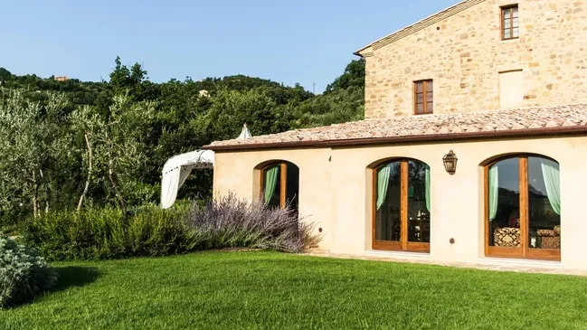 A scenic view of a rustic stone house with large windows, surrounded by lush greenery and trees, featuring a well-maintained lawn.