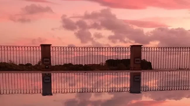 A serene sunset view with pink and orange hues reflected in a pool, featuring a decorative fence along the edge.