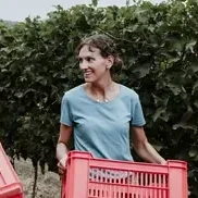 Two women smiling while carrying red harvest baskets in a vineyard.